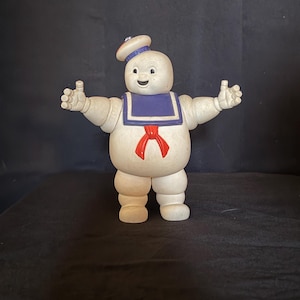 Puede incluir: Una figura blanca del hombre de malvavisco Stay Puft con un cuello de marinero azul, un pañuelo rojo y un sombrero de marinero blanco con una banda azul que lleva el texto Stay Puft. La figura tiene una expresión sonriente y los brazos extendidos.