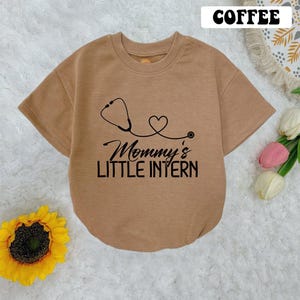 May include: A brown short-sleeved t-shirt with a black stethoscope and heart graphic. The text "Mommy's Little Intern" is printed on the shirt.