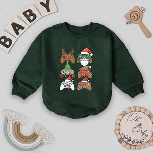 May include: Dark green baby bodysuit with six Christmas-themed video game controller designs. The controllers feature reindeer antlers, Santa hats, and Christmas trees. The word "BABY" is spelled out in wooden letters.