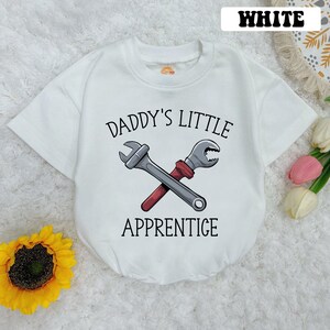 May include: White short-sleeved t-shirt with a graphic of two wrenches crossed over each other. The text "Daddy's Little Apprentice" is printed on the shirt.