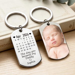 May include: Two silver-toned metal dog tag keychains. One features a printed calendar for April 2026 with the 16th circled, the text "The day you became my Daddy" and the name "Sophia". The other keychain displays a color photograph of a sleeping baby.