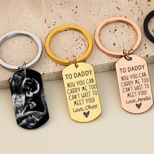 Op de afbeelding: Vier metalen sleutelhangers in de vorm van een dog tag in zilver, goud, roségoud en zwarte afwerking. De eerste toont een zwart-wit echografie. De andere zijn gegraveerd met: TO DADDY NOW YOU CAN CARRY ME TOO CAN'T WAIT TO MEET YOU!