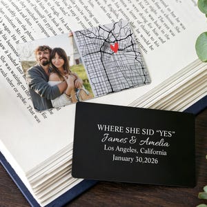 Pode incluir: Um cartão retangular de metal preto com texto gravado em branco que diz WHERE SHE SID "YES" James & Amelia Los Angeles, California January 30,2026, acompanhado por uma foto quadrada de um casal e um mapa com um coração vermelho.