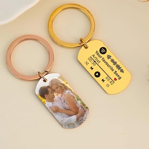 May include: Two gold keychains with a photo and a Spotify code. The keychain with the photo has a rose gold ring. The keychain with the Spotify code has a gold ring. The Spotify code says "Your favourite Song" with the artist and time displayed.