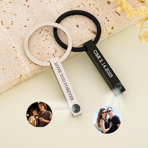 Könnte beinhalten: Zwei silberne Schlüsselanhänger mit schwarzen gravierten Balken. Ein Balken trägt die Aufschrift "Love You Forever" und der andere "C&K 2.14.2025". Jeder Balken hat ein kleines rundes Foto in das Ende eingebettet.