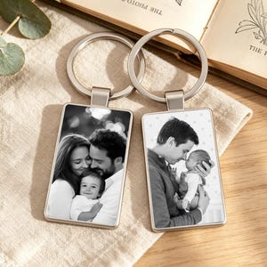 May include: Two rectangular metal keychains featuring black and white photographic prints. The left keychain displays a family of three, while the right keychain shows a person holding an infant. Both items include silver-toned metal split rings.