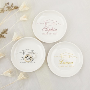 Op de afbeelding: Drie witte keramische ringenschalen met een afstudeerhoedontwerp. De schalen zijn gepersonaliseerd met de namen "Sophia", "Kelly" en "Lunna" en de tekst "Klas van 2024".