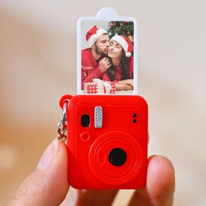 May include: A bright red miniature camera keychain featuring a pull-out rectangular photo insert. The photo displays a couple wearing Santa hats and red sweaters, with a Christmas tree and a gift box visible in the background of the image.