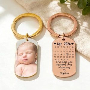Puede incluir: Dos llaveros personalizados. Uno presenta una foto de bebé, el otro tiene un calendario de abril de 2026 con el texto "The day you became my Mummy" y el nombre "Sophia". Ambos llaveros tienen anillos metálicos.