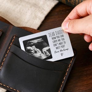 Pode incluir: Uma carteira bifold de couro castanho escuro com costuras visíveis e vários compartimentos para cartões. Um pequeno cartão retangular branco está inserido num compartimento, apresentando uma imagem de ecografia a preto e branco e o texto: Dear Daddy! Now you can carry me too. Can't wait to meet you! Love, the bump.