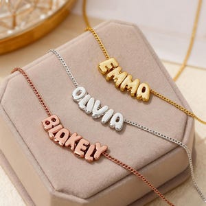 Könnte beinhalten: Drei personalisierte Namensketten in Gold, Silber und Roségold. Die Namen "Emma", "Olivia" und "Blakely" sind in einer Bubble-Letter-Schrift dargestellt. Jede Halskette hat eine zarte Kette und liegt auf einer hellrosa Oberfläche.