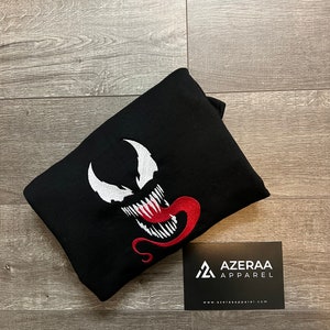 May include: Black hooded sweatshirt with a white embroidered Venom character with a red tongue. The sweatshirt is folded and a business card with the text "AZERAA APPAREL" and a website address is in the bottom right corner.
