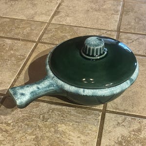 May include: A ceramic soup bowl with a lid and handle. The bowl is a deep teal green color with a light blue and white trim. The lid has a knob on top. The bowl is sitting on a brown tile surface.