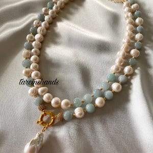 May include: A double-strand necklace featuring white pearls and light blue beads. The necklace has a gold clasp and a large, teardrop-shaped pearl pendant. The background is a soft, white fabric.