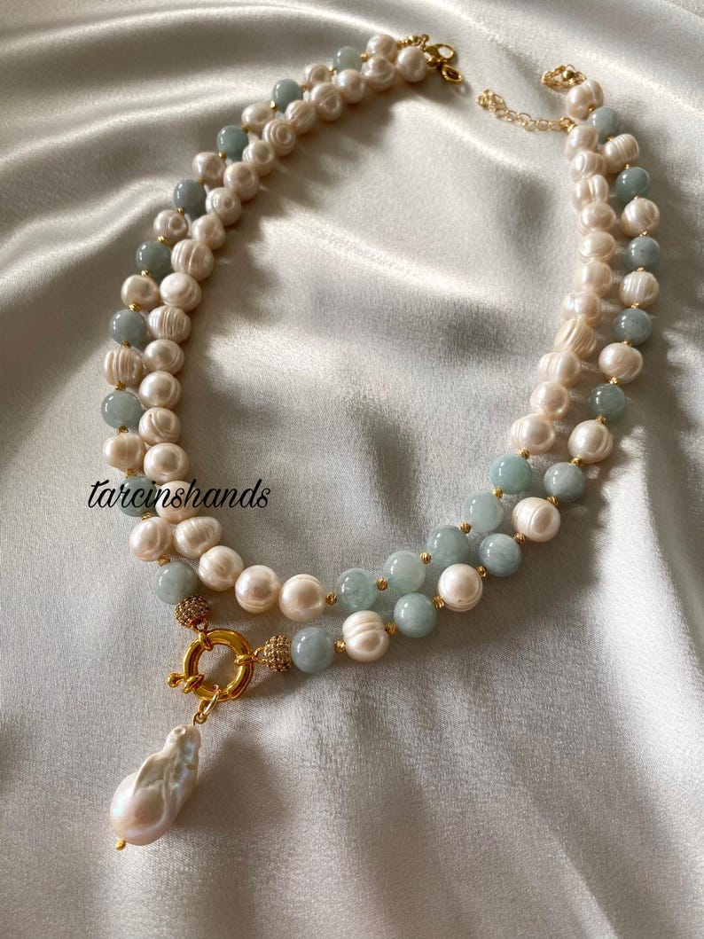 May include: A double-strand necklace featuring white pearls and light blue beads. The necklace has a gold clasp and a large, teardrop-shaped pearl pendant. The background is a soft, white fabric.