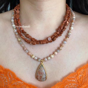 May include: Three layered necklaces featuring sunstone beads. The top layer has rough, reddish-brown beads, the middle layer has smaller, rounded beads, and the bottom layer has a teardrop pendant with a gold-colored frame. The necklaces are displayed on a person.