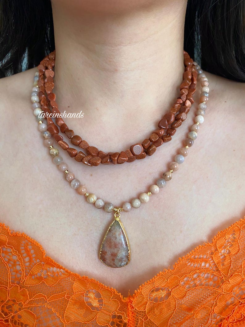 May include: Three layered necklaces featuring sunstone beads. The top layer has rough, reddish-brown beads, the middle layer has smaller, rounded beads, and the bottom layer has a teardrop pendant with a gold-colored frame. The necklaces are displayed on a person.
