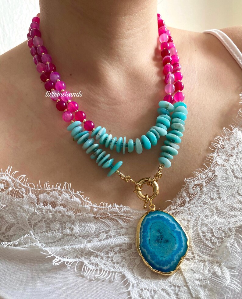 May include: A layered necklace with pink and turquoise beads and a large turquoise agate pendant. The necklace is on a white lace background.
