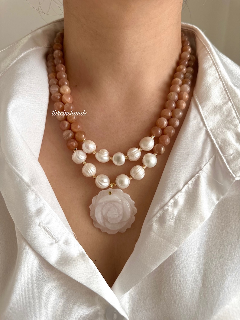 May include: A double-strand necklace with a rose-shaped pendant. The top strand is made of light brown sunstone beads, and the bottom strand is made of white pearls. The pendant is made of white jade and has a delicate rose design.