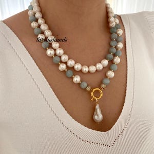 May include: A layered necklace featuring white pearls, light blue beads, and a gold-colored clasp with a pear-shaped pearl pendant. The necklace is worn against a white top.