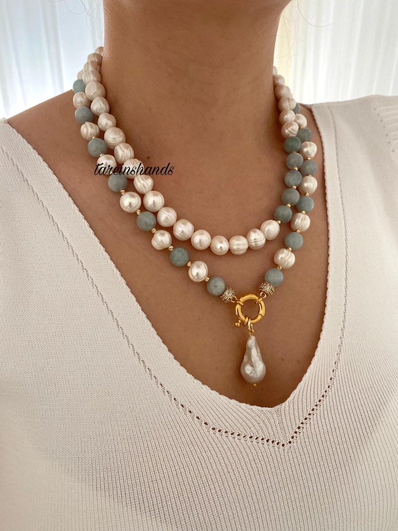 May include: A layered necklace featuring white pearls, light blue beads, and a gold-colored clasp with a pear-shaped pearl pendant. The necklace is worn against a white top.