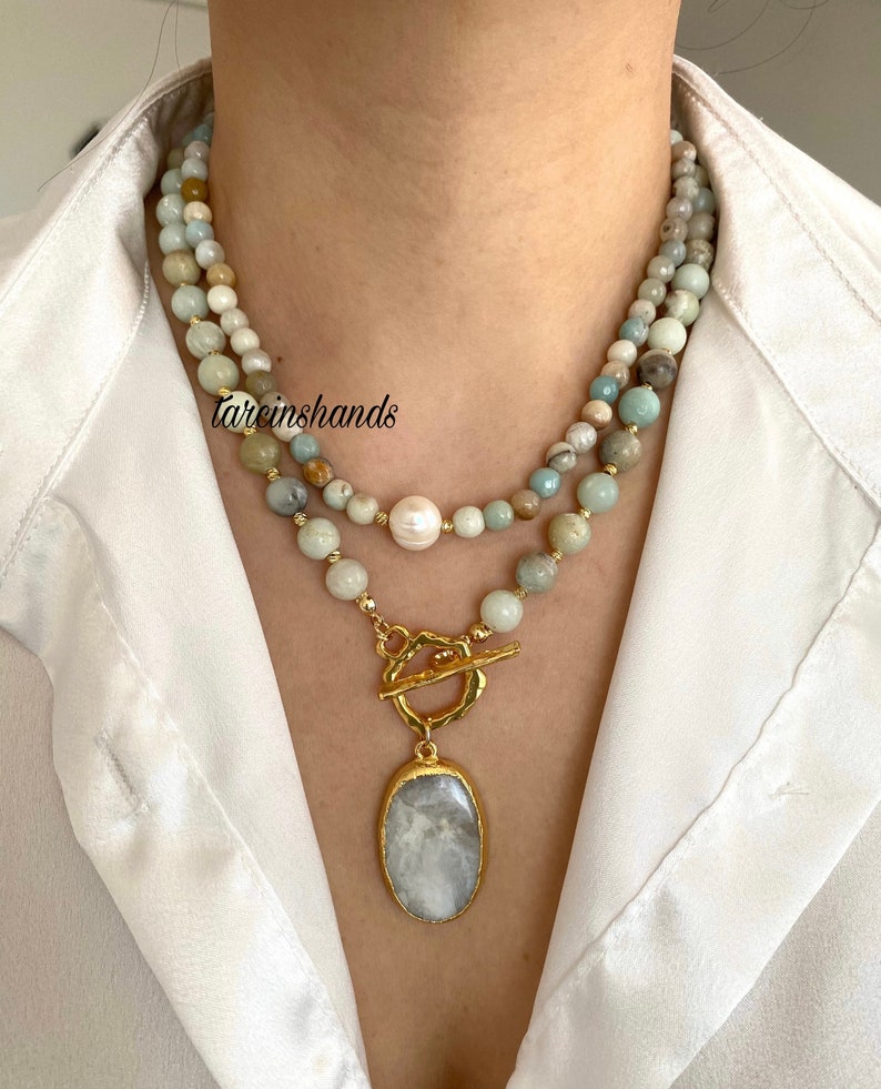 Puede incluir: Un collar de varias capas con un cierre de palanca dorado y un gran colgante ovalado. El collar est&aacute; hecho de cuentas de color verde claro, marr&oacute;n y blanco. El colgante est&aacute; hecho de una piedra gris claro.