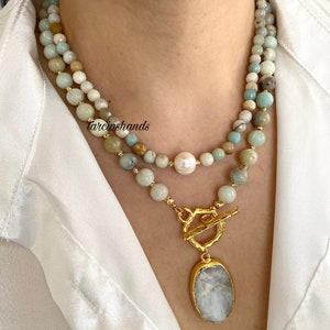 Puede incluir: Un collar de varias capas con un colgante dorado que presenta una piedra blanca ovalada grande. El collar est&aacute; hecho de cuentas de color verde claro y blanco con una sola perla blanca.