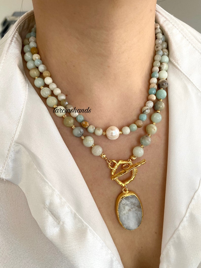 Puede incluir: Un collar de varias capas con un colgante dorado que presenta una piedra blanca ovalada grande. El collar est&aacute; hecho de cuentas de color verde claro y blanco con una sola perla blanca.