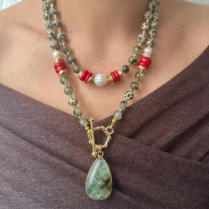 May include: A layered necklace featuring a gold clasp and a teardrop-shaped pendant with green and brown inclusions. The necklace includes speckled beads, red cylindrical beads, and a pearl. The necklace is worn against a brown background.