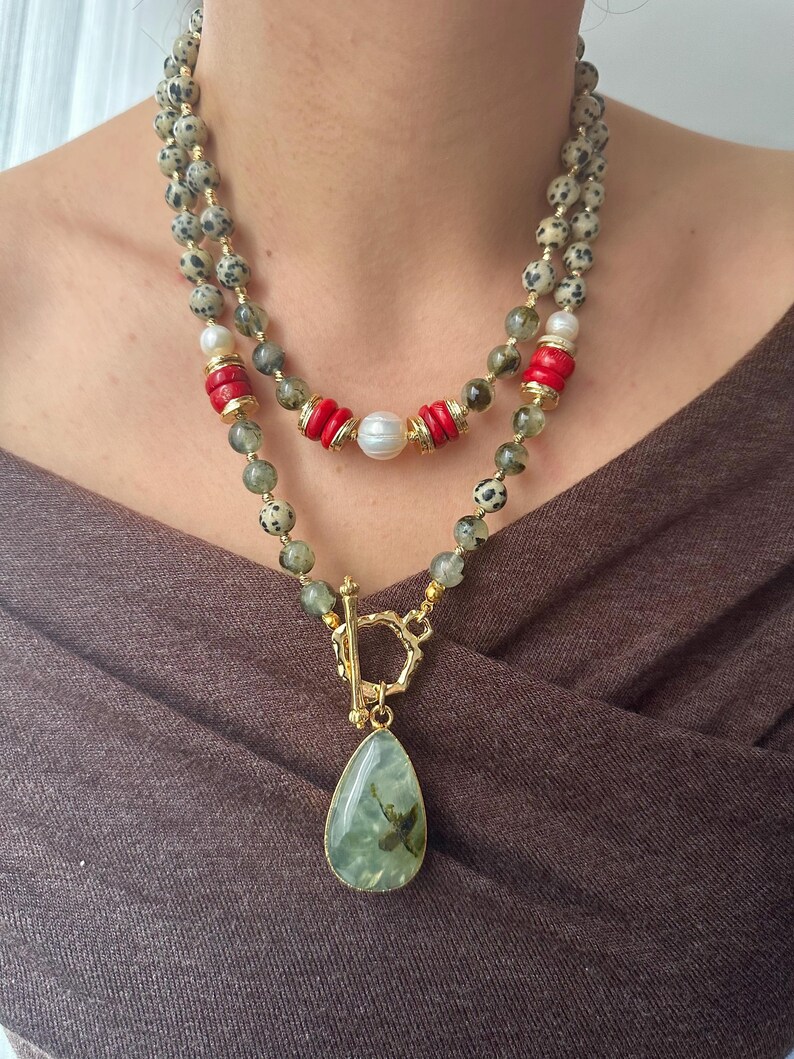 May include: A layered necklace featuring a gold clasp and a teardrop-shaped pendant with green and brown inclusions. The necklace includes speckled beads, red cylindrical beads, and a pearl. The necklace is worn against a brown background.