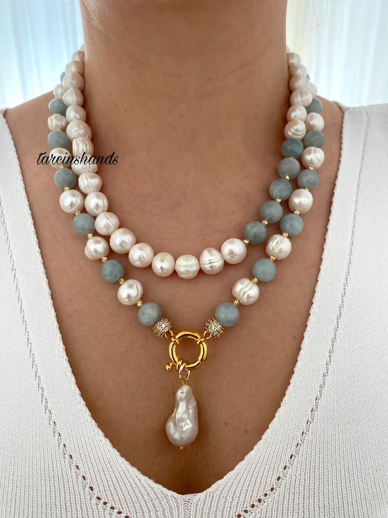 May include: A two-strand necklace featuring white and light blue beads, with a gold-colored clasp and a large, irregular white pearl pendant. The necklace is worn around the neck, showcasing the jewelry's design and color contrast.