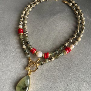 May include: A layered necklace featuring speckled beads, red cylindrical beads, and a pear-shaped green pendant with a gold-colored frame. The necklace includes a gold-colored clasp and white pearl beads.