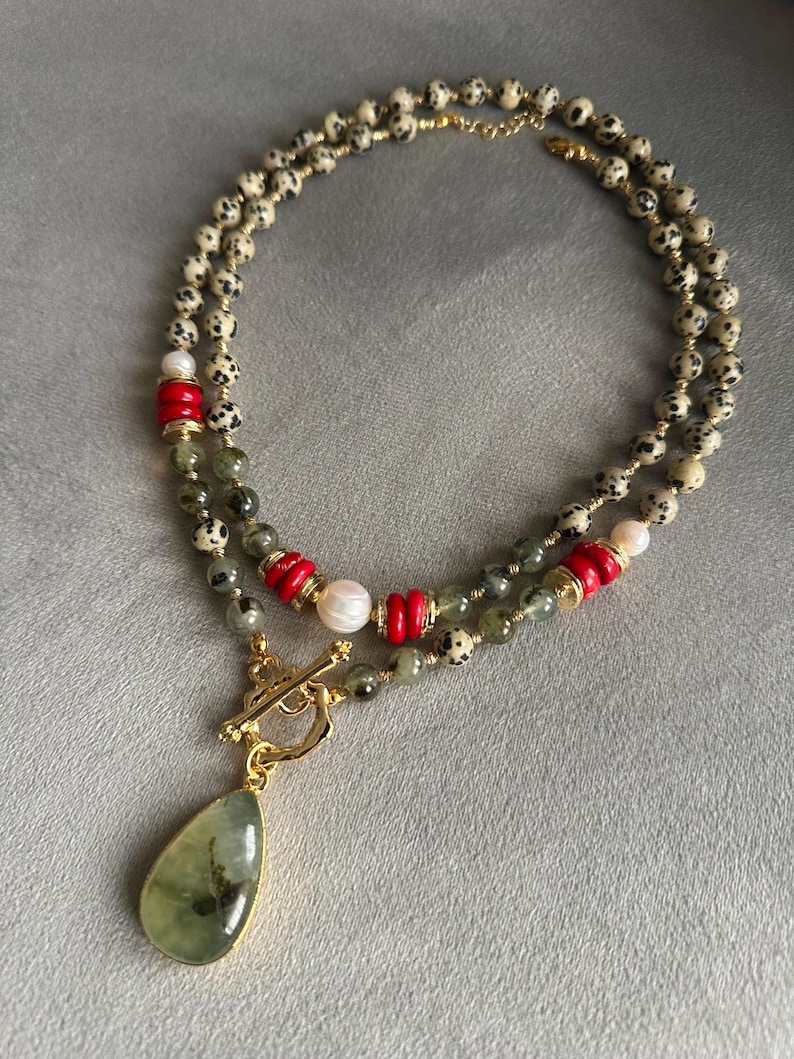 May include: A layered necklace featuring speckled beads, red cylindrical beads, and a pear-shaped green pendant with a gold-colored frame. The necklace includes a gold-colored clasp and white pearl beads.
