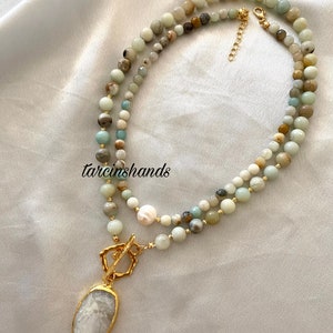 Puede incluir: Un collar de m&uacute;ltiples hilos con cuentas verdes, blancas y marrones. El collar tiene un cierre dorado y un colgante grande de forma ovalada con una piedra blanca.