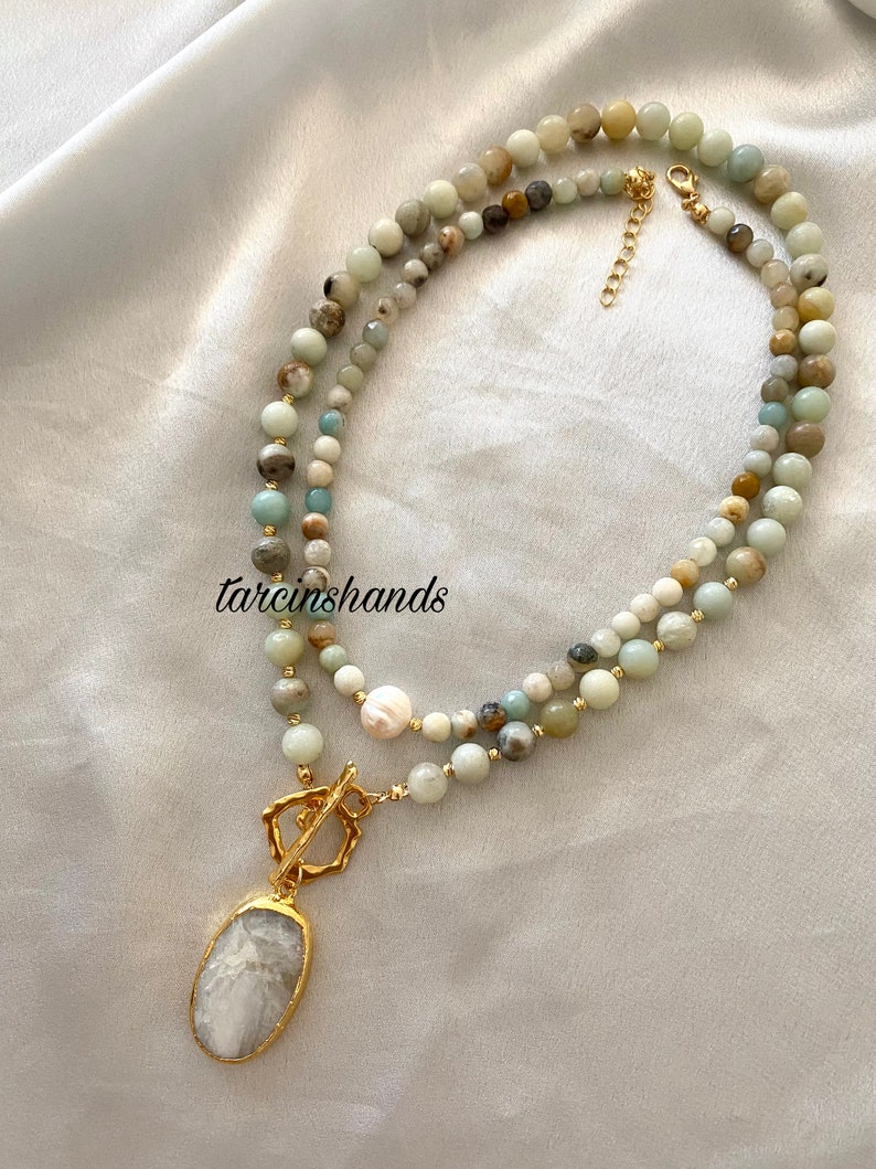Puede incluir: Un collar de m&uacute;ltiples hilos con cuentas verdes, blancas y marrones. El collar tiene un cierre dorado y un colgante grande de forma ovalada con una piedra blanca.