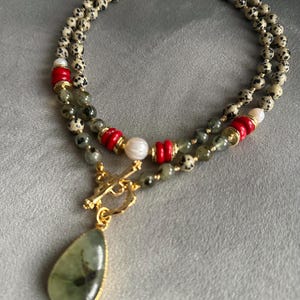 May include: A layered necklace featuring a teardrop-shaped green pendant with a gold-colored frame. The necklace includes speckled beads, red and gold accents, and a gold-colored clasp. The design incorporates a mix of textures and colors.