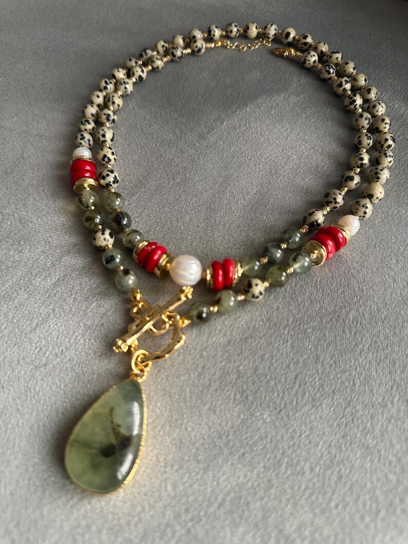 May include: A layered necklace featuring a teardrop-shaped green pendant with a gold-colored frame. The necklace includes speckled beads, red and gold accents, and a gold-colored clasp. The design incorporates a mix of textures and colors.