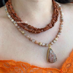 May include: A layered necklace featuring three strands of gemstones. The top strand is made of rough, reddish-brown stones, the middle strand is a mix of beige and brown beads, and the bottom strand has a teardrop pendant.