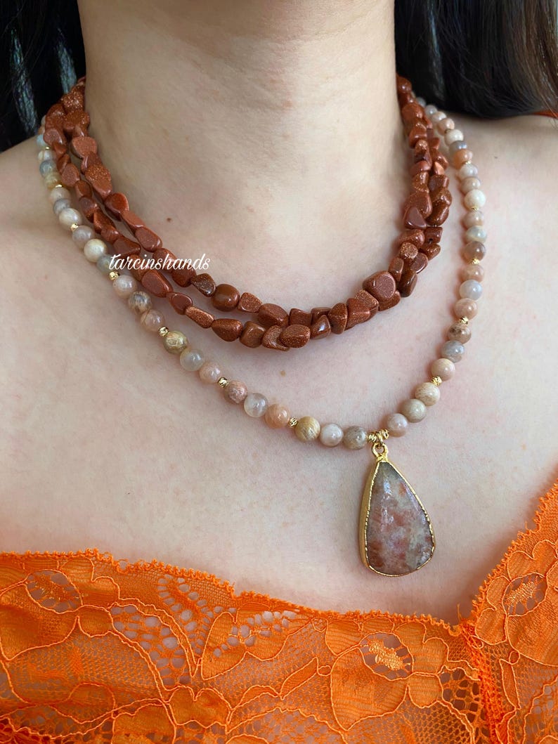 May include: A layered necklace featuring three strands of gemstones. The top strand is made of rough, reddish-brown stones, the middle strand is a mix of beige and brown beads, and the bottom strand has a teardrop pendant.