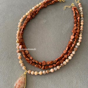May include: A multi-strand necklace featuring three layers of beads in shades of brown, tan, and gold. The necklace includes a teardrop-shaped pendant with a gold-colored frame. The text "tarcinshands" is visible on the image.
