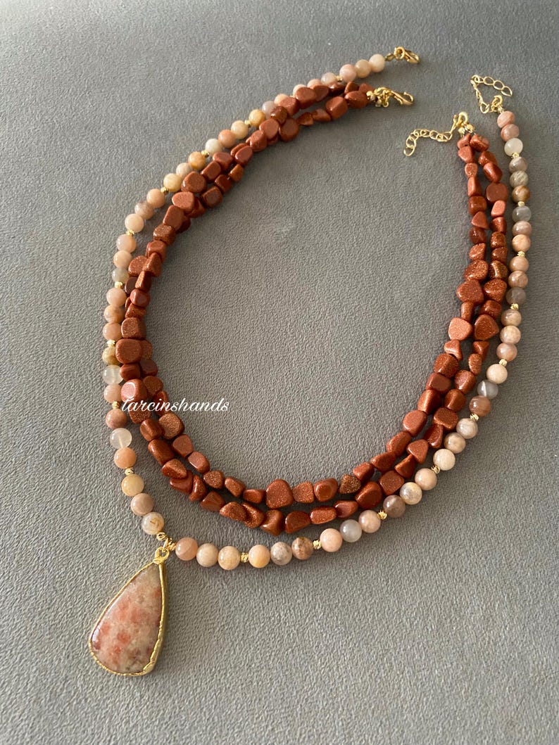 May include: A multi-strand necklace featuring three layers of beads in shades of brown, tan, and gold. The necklace includes a teardrop-shaped pendant with a gold-colored frame. The text "tarcinshands" is visible on the image.
