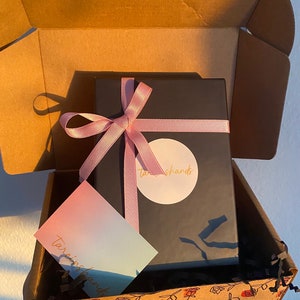 May include: A black gift box with a pink ribbon and a white circle sticker with the text "tarishards" on it. The box is inside a brown cardboard box with a rose pattern on the bottom.