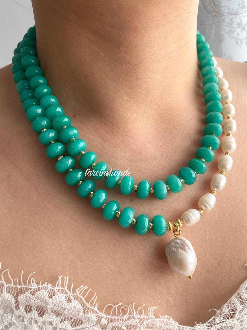 Puede incluir: Un collar de varias capas con cuentas de color turquesa y perlas blancas. El collar tiene detalles dorados y un colgante de perla grande de forma irregular. El texto "tarcinshands" es visible en el collar.