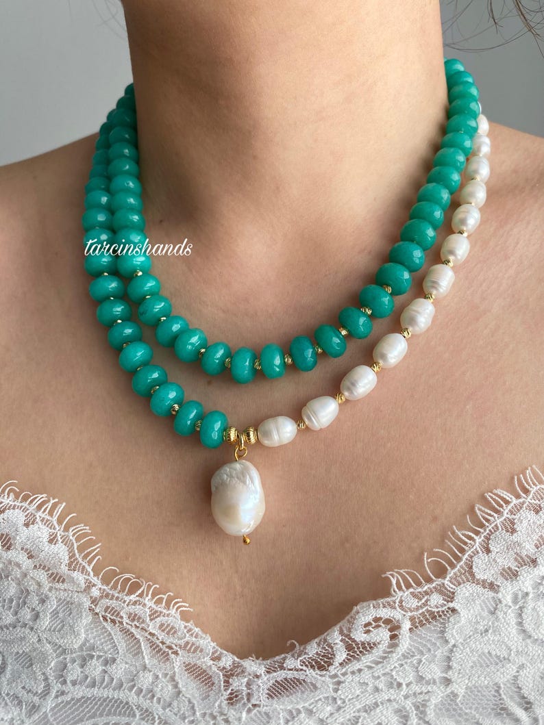Puede incluir: Un collar de dos hilos con cuentas turquesas y perlas blancas. Las cuentas turquesas est&aacute;n ensartadas con detalles dorados. Un gran colgante de perla barroca blanca cuelga del collar. El texto "tarcinshands" es visible.