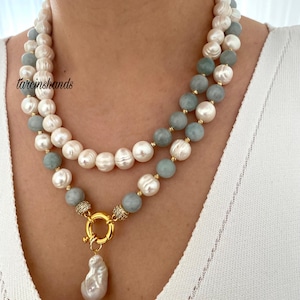 May include: A two-strand necklace featuring white and light blue beads, with a gold clasp and a large, irregular pearl pendant. The necklace is worn against a white top. The text "tarcinshands" is visible.
