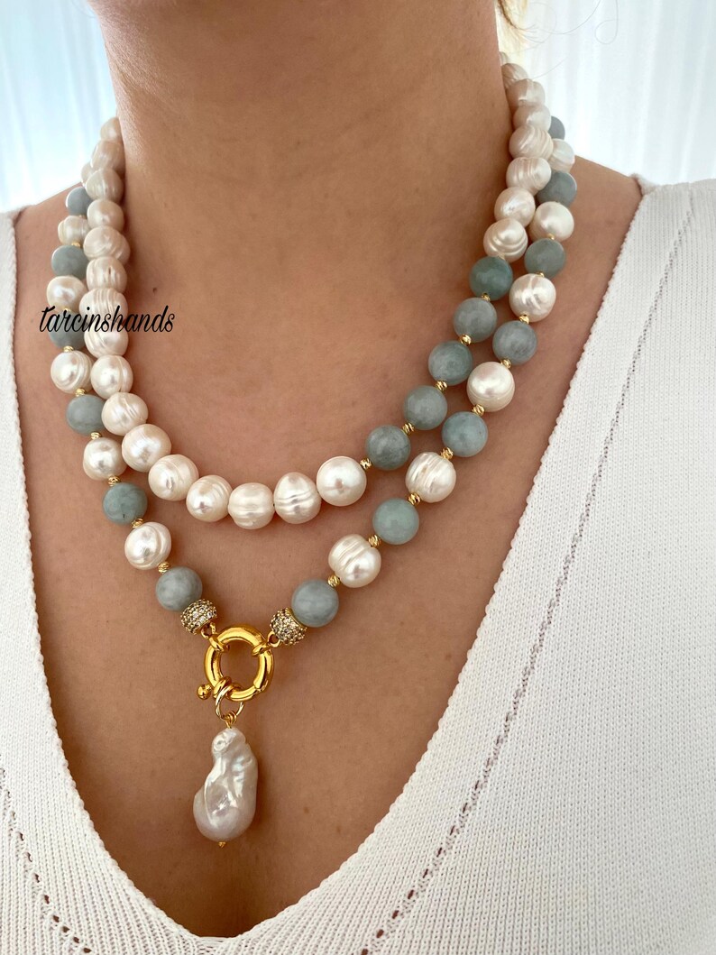 May include: A two-strand necklace featuring white and light blue beads, with a gold clasp and a large, irregular pearl pendant. The necklace is worn against a white top. The text "tarcinshands" is visible.