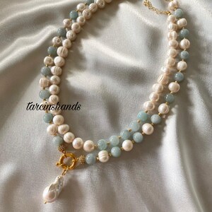 May include: A layered necklace featuring white pearls and light blue beads. The necklace has a gold clasp and a large, pear-shaped pearl pendant. The background is a white, silky fabric. The text "tarcinshands" is in the upper left corner.
