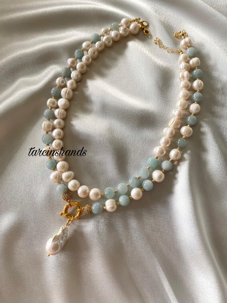 May include: A layered necklace featuring white pearls and light blue beads. The necklace has a gold clasp and a large, pear-shaped pearl pendant. The background is a white, silky fabric. The text "tarcinshands" is in the upper left corner.