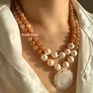 May include: A double-strand necklace with peach-colored sunstone beads and white pearls. The necklace has a white carved rose pendant.