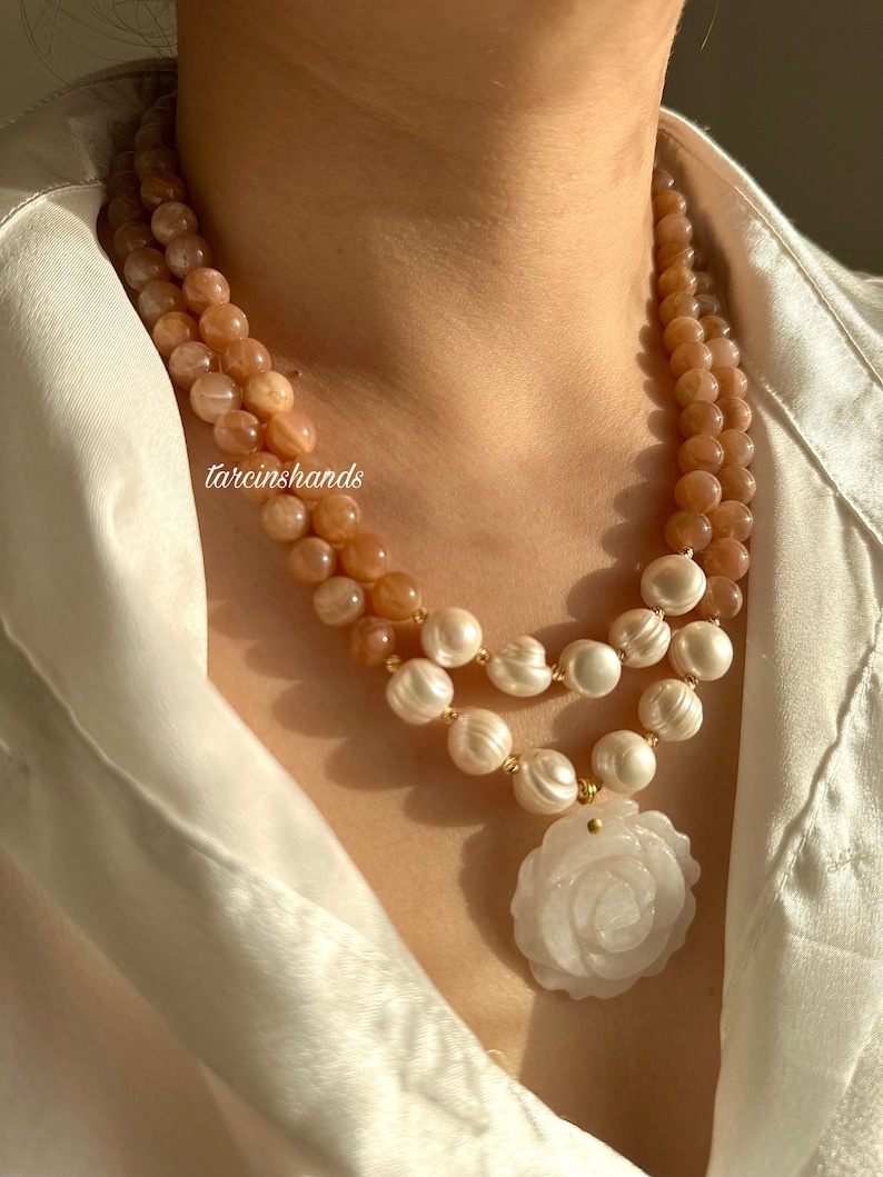 May include: A double-strand necklace with peach-colored sunstone beads and white pearls. The necklace has a white carved rose pendant.